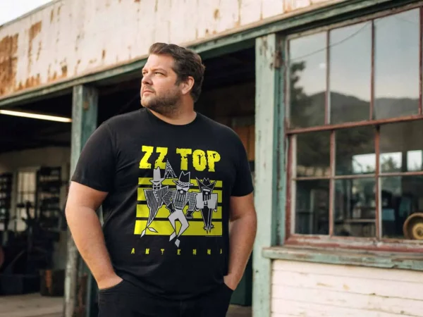 A man wearing a ZZ Top concert tee standing outdoors in front of an old, weathered building with rustic charm and large windows, emphasizing retro and rock style.
