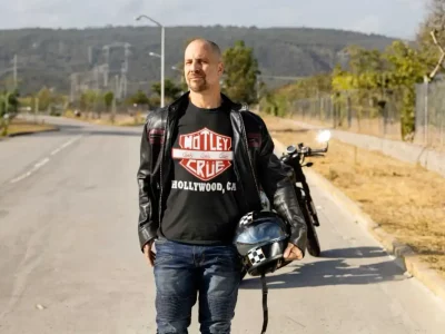 Man in a black leather jacket wearing a Mötley Crüe “Hollywood, CA” t-shirt stands on an open road holding a motorcycle helmet, with a motorcycle behind him.