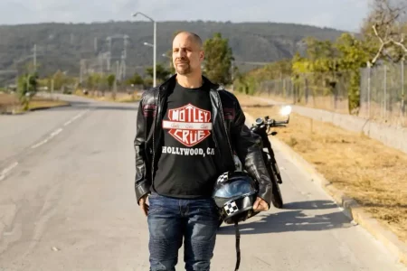 Man in a black leather jacket wearing a Mötley Crüe “Hollywood, CA” t-shirt stands on an open road holding a motorcycle helmet, with a motorcycle behind him.