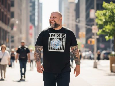 Bold man in graphic T-shirt walking through busy city street with diverse pedestrians and bright sunlight, showcasing urban fashion and vibrant city life.