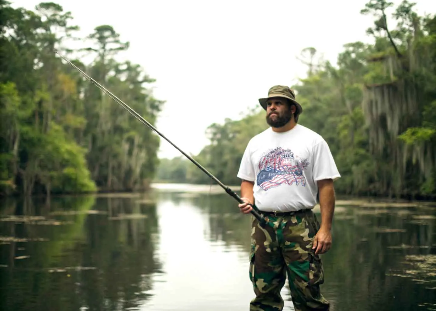 Alt text: Man fishing in a river surrounded by lush trees, wearing a hat and camouflage pants.