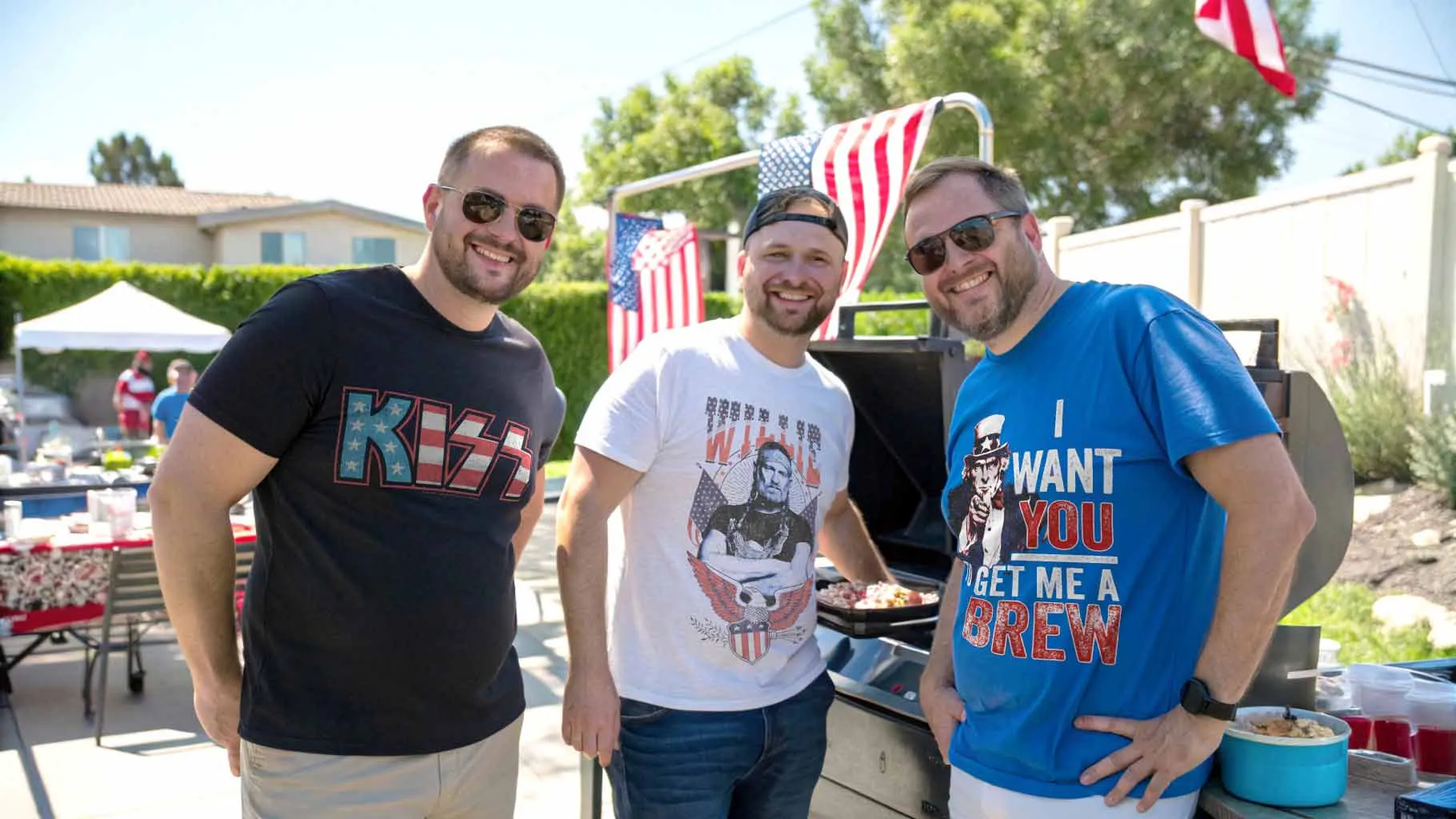 Men smiling in casual T-shirts with patriotic and humor designs at outdoor party.