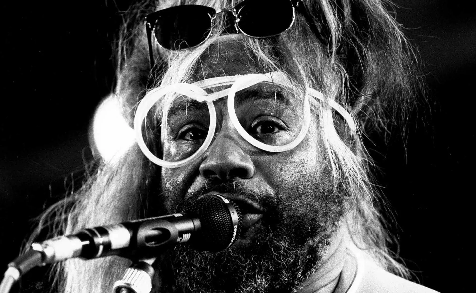 Close-up of a fun, expressive person with white hair and glasses singing into a microphone.