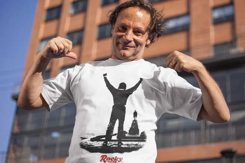 Man wearing a Rocky-themed T-shirt outdoors, flexing muscles in front of a building.