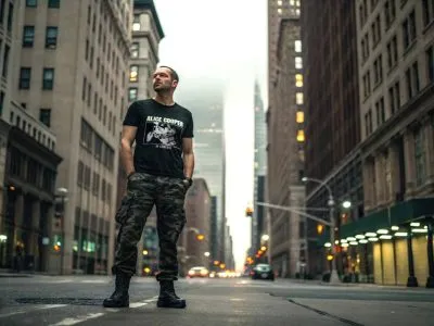 A man stands confidently in the middle of a city street during evening, wearing a black graphic t-shirt and camouflage pants, showcasing urban fashion style.