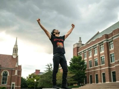A person celebrating outdoors on a college campus with raised fists, symbolizing victory and empowerment, in front of historic brick buildings.
