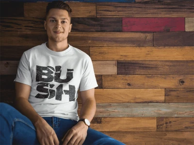 Smiling young man wearing a "BUSH" graphic tee, sitting against a rustic wooden wall, showcasing trendy casual fashion and urban style.