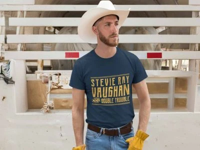 A man in a cowboy hat and graphic tee with "Stevie Ray Vaughan Double Trouble" stands against a rustic fence with cows in the background.