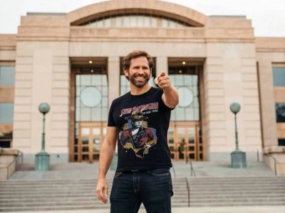 A smiling man pointing forward in front of a grand courthouse building, wearing a graphic T-shirt, representing confidence and style.