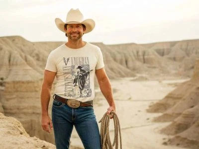 A smiling man wearing a cowboy hat and graphic t-shirt stands in a desert canyon holding a lasso, showcasing western-inspired fashion and outdoor adventure style.