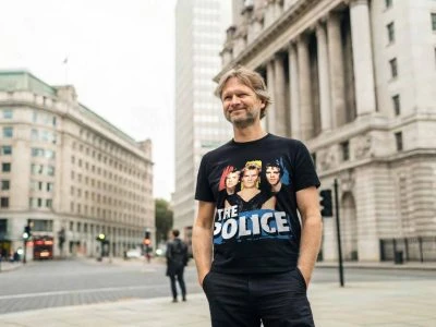 A man stands confidently on a city street wearing a vintage The Police band T-shirt, surrounded by historic architecture, capturing a casual urban vibe.