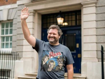 A man with a beard cheers and waves during a casual outdoor event, wearing a graphic T-shirt featuring the band The Police, in front of a historic building.