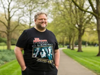 A smiling man wearing a black graphic T-shirt with Japanese text, standing on a tree-lined park path in spring.