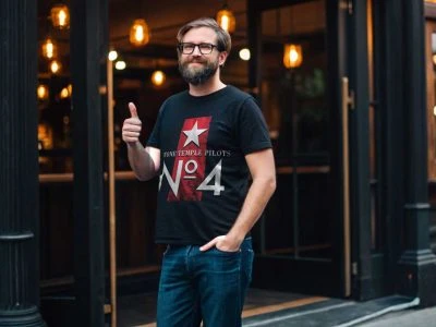 Thrilled man wearing a Rock Band T-Shirt shows positivity outside a stylish bar or restaurant. Perfect for music lovers and casual fashion fans.
