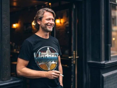 Relaxed man wearing a "WARRAN" graphic t-shirt, standing outside a trendy bar or cafe at night.