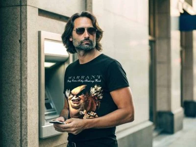 Stylish man with sunglasses using ATM in urban setting, wearing graphic T-shirt featuring bold artwork, showcasing casual fashion and street style.