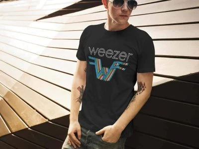 A young man wearing a Black Weezer band graphic t-shirt, sunglasses, and jeans, leaning against modern wooden architecture in an outdoor setting.